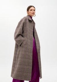 Ruudullinen, ylipitkä takki ruskean, valkoisen ja purppuran sävyissä, jossa on kaulus, leveät hihat ja sivutaskut, päällä purppura paita.