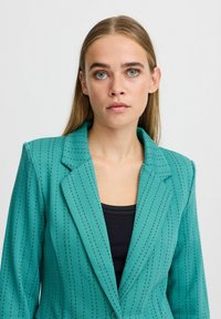 Blazer turchese con motivo a righe sottili, revers strutturato, chiusura a un bottone e texture liscia. Indossato sopra un top nero.
