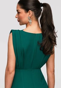 Femme avec une longue queue de cheval foncée portant une robe verte sans manches et de grandes boucles d'oreilles en argent en forme de fleur, vue de dos et de profil.