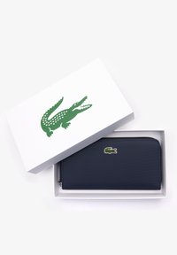 Portefeuille zippé bleu marine avec surface texturée, présentant un logo de crocodile vert, présenté dans une boîte blanche avec un design de crocodile.