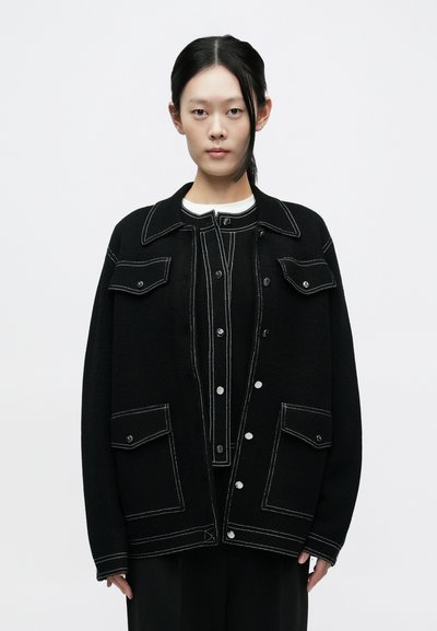 Giacca nera oversize con cuciture bianche a contrasto, caratterizzata da grandi tasche, colletto a punta e bottoni a pressione. Tessuto spesso e testurizzato.