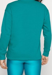 Turkusowy sweatshirt z okrągłym dekoltem, długimi rękawami i ściągaczami, w połączeniu z błyszczącymi turkusowymi legginsami. Gładka tekstura materiału.
