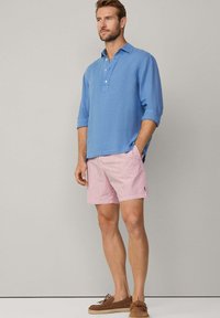 Hellblaues Leinenhemd mit Knopfleiste und hochgerollten Ärmeln, kombiniert mit rot-weißen gestreiften Shorts und braunen Bootsschuhen.
