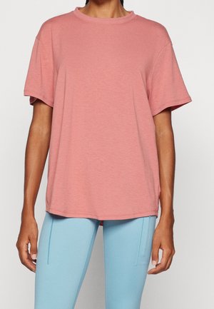 Vrouw die een losse roze shirt met korte mouwen en lichtblauwe nauwsluitende broek draagt, staand tegen een effen witte achtergrond.