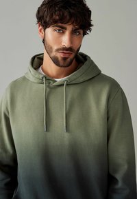 Zeleni hoodie s gradijentnim uzorkom, prednjim kengur džepom, kapuljačom na vezanje i teksturiranom tkaninom. Sadrži srebrne metalne dijelove na vezicama.