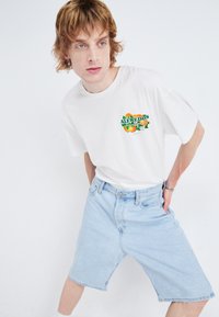 Hvit T-skjorte med fargerik "ALFREDO'S" grafikk og lyseblå denimshorts. Shortsen er kne-lang med en avslappet passform og sydde lommer.