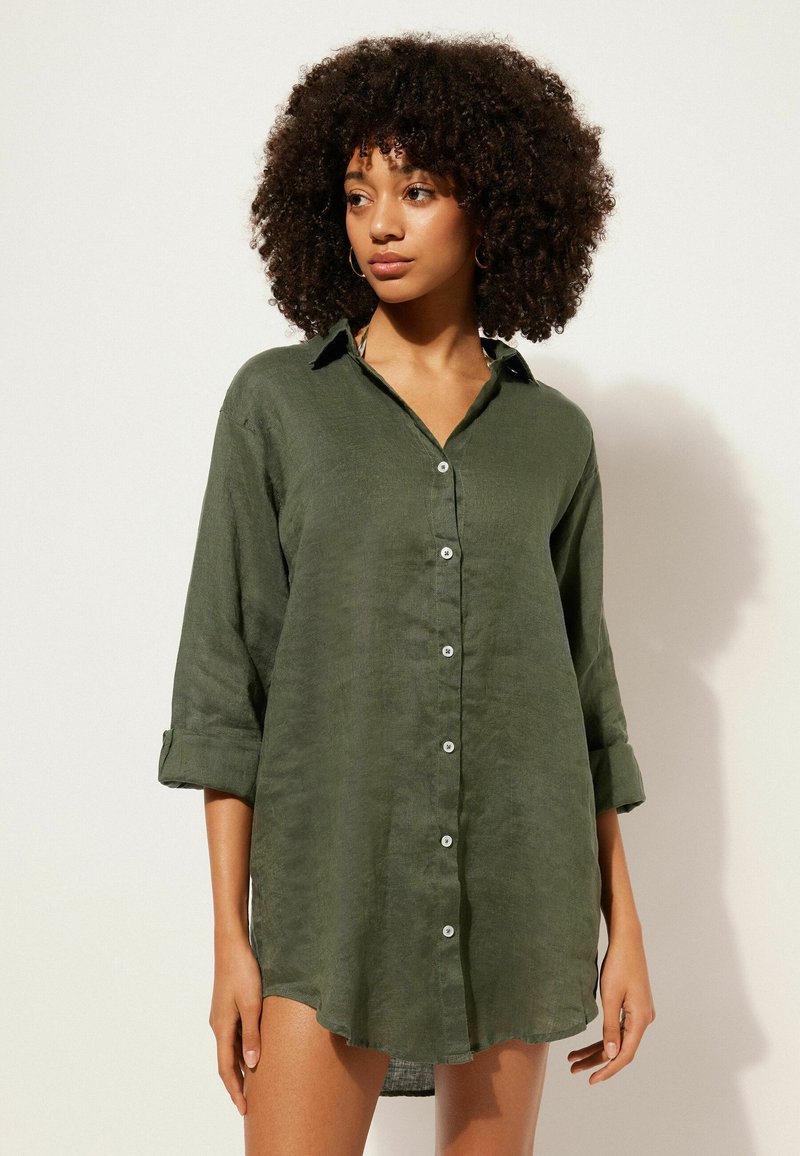 Camicia botton-down verde scuro in tessuto leggero, con colletto, maniche lunghe arrotolate e orlo curvo. I dettagli dei bottoni sono visibili.