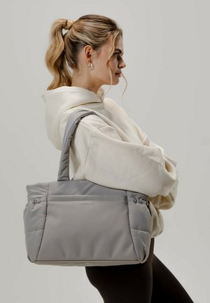 Sac tote matelassé gris avec des accents lisses, des poignées rembourrées et une poche zippée à l'avant. Le mannequin tient le sac contre un arrière-plan neutre.