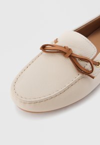 Μπεζ δερμάτινο μοκασίνι με ραμμένες λεπτομέρειες και καφέ δερμάτινο φιόγκο δεμένο στην κορυφή, εμφανιζόμενο σε λευκό φόντο.