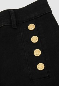 Tissu noir avec quatre boutons dorés verticaux embossés d'un motif floral et une passoire de ceinture visible sur le côté gauche.