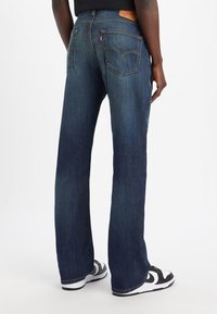 Donkerblauwe denim jeans met een rechte pasvorm, voorzien van achterzakken en een leren label met merknaam op de taille. Gemaakt in combinatie met zwart-witte sneakers.