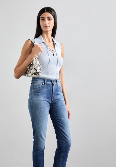 Top sin mangas de canalé azul claro con detalles de cordones, combinado con jeans ajustados azules; sostiene un pequeño bolso de piel de serpiente.