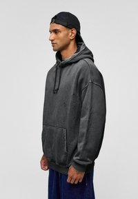 Grijze, oversized hoodie gemaakt van zachte stof. Heeft een voorzak, een capuchon met koord en een iets versleten textuur voor extra detail.