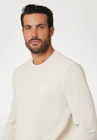 Krämfärgad crewneck-tröja i strukturerad tyg, med en liten vit logotyp på vänster bröst. Enkel, figursydd design.