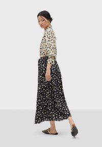 Blusa beige con pois neri, maniche lunghe, abbinata a una gonna nera con un motivo simile. Scarpe nere slip-on con borchie.