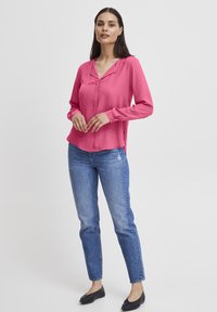 Long-sleeve, pink blouse with a v-neck and button detailing, paired with light blue denim jeans and black flats. Simple and modern design.