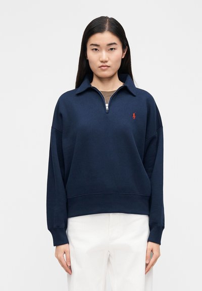 Tumesinist värvi pullover kapuutsiga, lukuga kaeluse ja soonikuga varrukasäärtega ning väike punane logo rinnal. Pehme kangas ja mugav lõige.