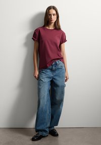 T-shirt corto color bordeaux abbinato a jeans larghi e sbiaditi di colore blu, e scarpe nere. L'outfit presenta maniche arrotolate e una vestibilità comoda.