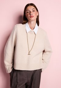 Young woman with straight brown hair wearing a beige knit sweater over a white collared shirt and brown pinstriped trousers, standing against pink wall.