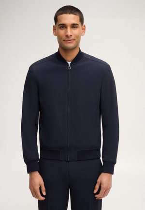 Uomo che indossa una giacca bomber blu navy con zip, colletto, polsini e orlo a costine, in piedi davanti a uno sfondo chiaro e uniforme.