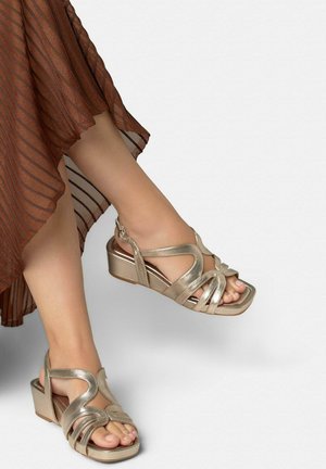 Gambe con sandali bassi color oro metallizzato con più cinturini e fibbia alla caviglia, abbinati a una gonna plissettata marrone.