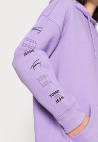 Lilla hoodie med sorte trykte logoer på begge ærmer. Lavet af blødt materiale med en afslappet pasform og hætte med snøre.