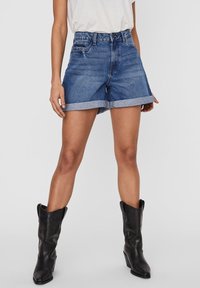 Kvinna i rullade denimshorts, vit topp och svarta westernstövlar vid vadleden står mot en neutral bakgrund.