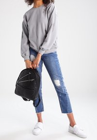Gray sweatshirt with raglan sleeves and minimal text, paired with distressed blue jeans and white slip-on shoes. Black backpack with zippers.