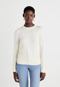 Pull en tricot de couleur crème avec un col rond et des manches longues. Présente des accents de boutons sur les ourlets des manches et un ourlet côtelé.
