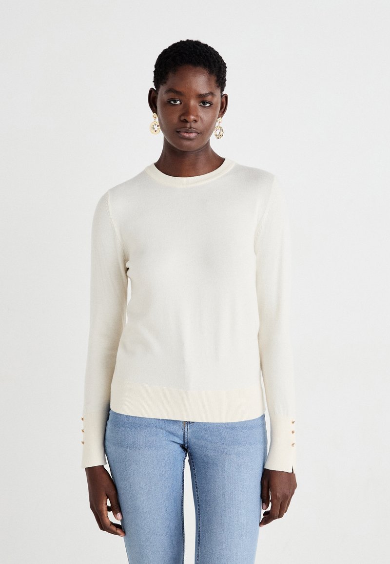 Pull en tricot de couleur crème avec un col rond et des manches longues. Présente des accents de boutons sur les ourlets des manches et un ourlet côtelé.