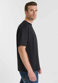 T-shirt nera a maniche corte con scollatura rotonda, realizzata in tessuto liscio. Il modello la indossa con jeans blu, esibendo una vestibilità semplice e dritta.