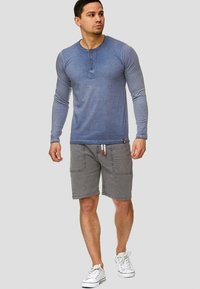 Chemise henley bleue à manches longues avec patte de boutonnage à trois boutons, associée à un short gris clair avec poches avant et cordon de serrage, portée avec des baskets blanches.