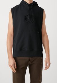 Bezrękawnik w kolorze czarnym z kapturem, z przednią kieszenią oraz wytłoczonym logo, noszony z brązowymi spodniami, przedstawiony od ramion do bioder.