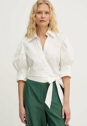 Donna bionda con camicetta bianca a portafoglio con maniche a sbuffo e pantaloni verdi a vita alta, in piedi contro uno sfondo chiaro e uniforme.