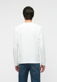 Bela majica s dolgimi rokavi iz bombaža, z okroglim izrezom in brezšivnimi rameni, v kombinaciji z modrimi jeans hlačami.