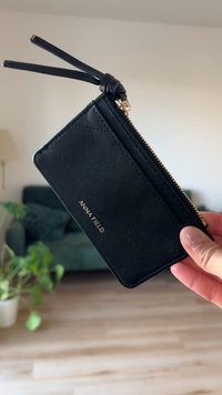 Portofel din piele neagră pentru carduri, cu un fermoar auriu, cu numele de marcă "ANNA FIELD" embosat și un design rectangular elegant, cu un finisaj texturat.
