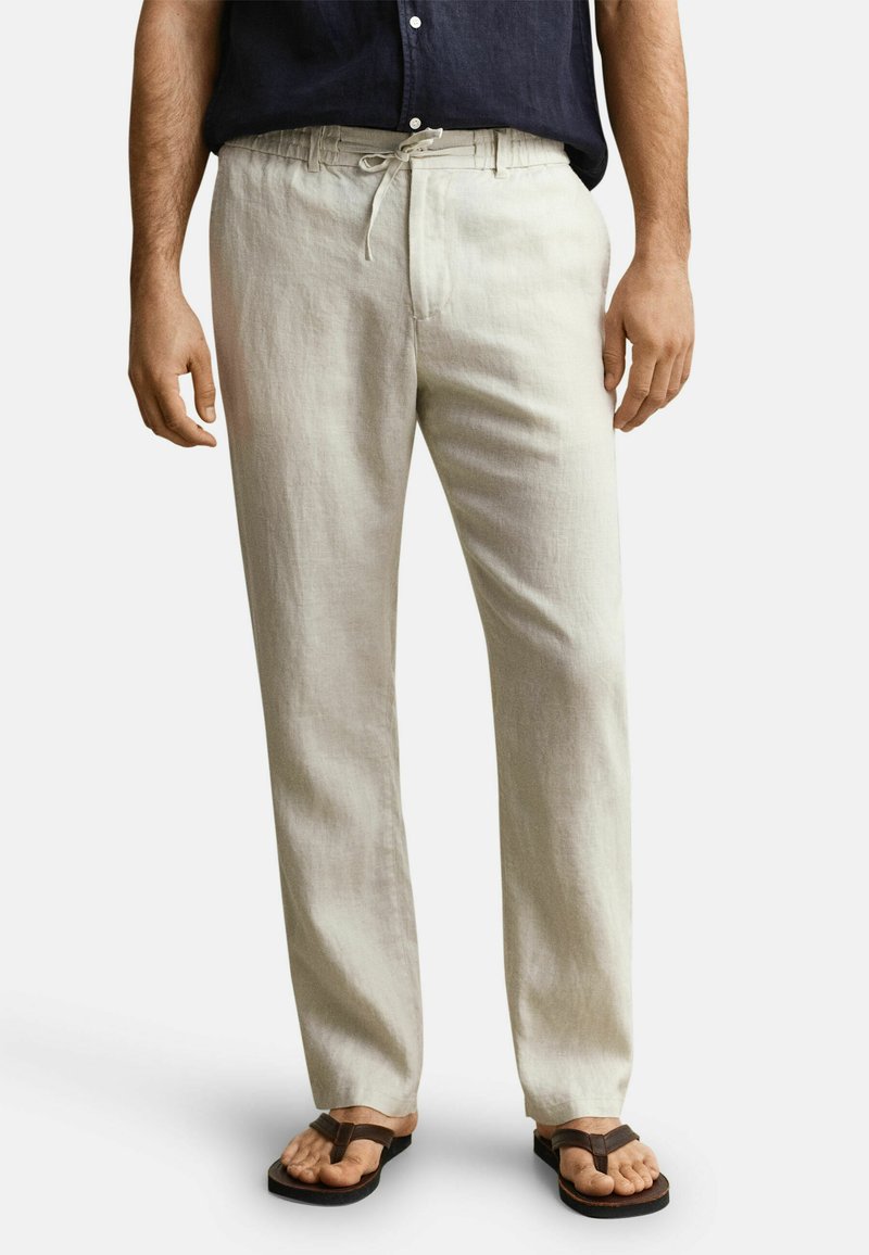 Man draagt beige linnen broek met trekkoord in de taille, donkerblauw overhemd en bruine slippers, staand tegen een effen achtergrond.