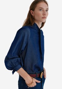 Navyblauwe blouse van zachte stof, met lange pofmouwen, staande kraag en een strikdetail bij de hals, gecombineerd met een riem en jeans.