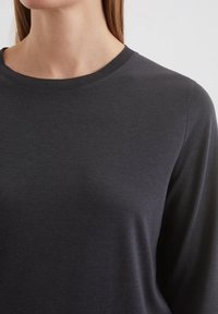 Žena oblečená v tmavě šedém dlouhém rukávu s žebrovaným kulatým výstřihem, zobrazená od krku po horní část hrudníku.