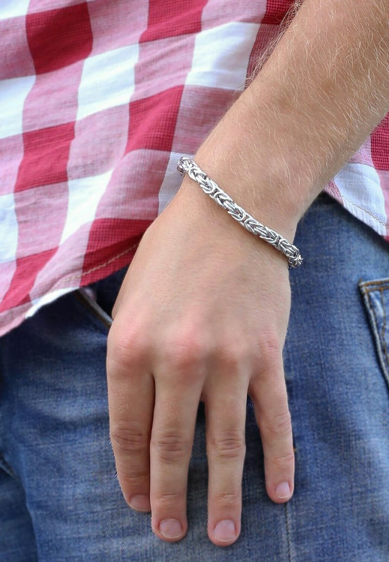 Silver braided bracelet on a light-skinned hand, with a checkerboard patterned shirt in the background. Material appears shiny and textured.