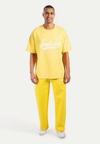 Camiseta grande de algodón amarilla con texto blanco y pantalones deportivos amarillos a juego, con un ajuste holgado y sutiles detalles de logo.