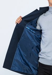 Marinejas met een wollen buitenkant, voorzien van een blauwe satijnen voering en een klein binnenzakje met een decoratieve rand. De zoom heeft contrasterende stiksels.