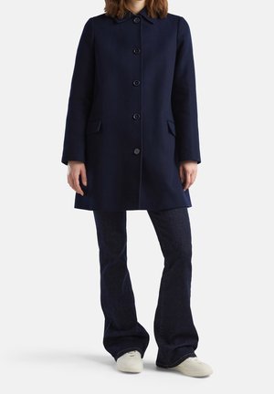 Personne portant un manteau bleu marine foncé boutonné, un jean évasé foncé et des baskets blanches, debout devant un fond clair uni.