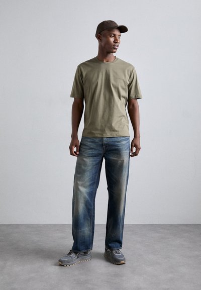 T-shirt en coton vert olive, jeans droits bleu déchirés, chaussures de sport grises et casquette marron, se tenant devant un fond neutre.