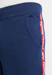 Pantalons de survêtement bleu marine avec une taille côtelée et une poche latérale. Présentent une bande rouge avec un texte blanc indiquant "ENLEVER AVANT LE VOL."