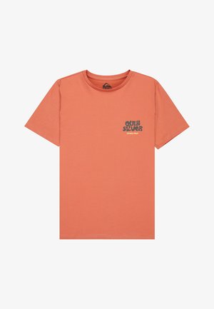 Garçon debout portant un t-shirt rose saumon avec un petit motif, un pantalon vert et des baskets marron à lacets blancs sur un fond uni.