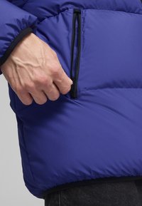 Lila pufferejacket sima, fényes textúrával; cipzáras zsebbe és fekete cipzárra van ellátva. Az ujjak sötét anyaggal vannak díszítve.