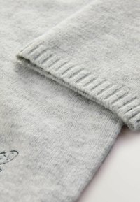 Dettaglio di un maglione grigio chiaro con una morbida texture lavorata a maglia, orlo a coste e polsini ripiegati, caratterizzato da leggere variazioni nel colore del filato.