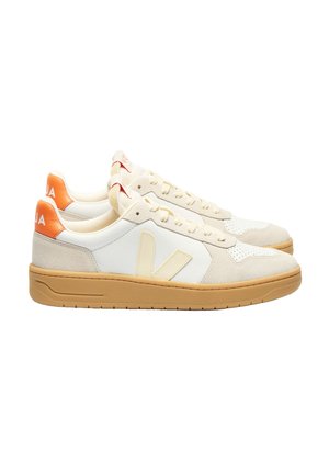 Zapatillas beige con parte superior de cuero blanco y detalles de ante, que presentan una lengüeta trasera naranja y una suela de goma marrón. Diseño de puntera perforada.
