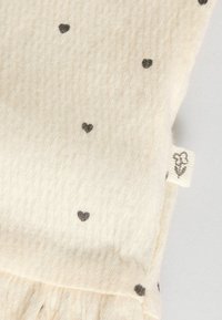 Tela de color crema con una textura suave, que presenta pequeños patrones de corazones oscuros. Incluye una etiqueta con un diseño floral en un lado.